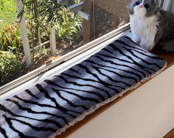 Puede incluir: Un cojín rectangular para el asiento de la ventana con un estampado de rayas de cebra grises y blancas. El cojín tiene una textura suave y afelpada. Un juguete de peluche con forma de gato está en el alféizar de la ventana.