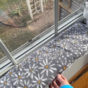 May include: A rectangular window perch cushion featuring a soft gray fleece fabric with a repeating white daisy pattern and yellow centers. The cushion has a white textured underside and is positioned along the base of a window frame.