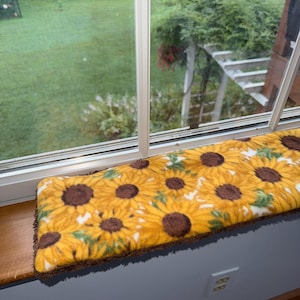 May include: A rectangular window perch cushion featuring a vibrant sunflower pattern with yellow petals and dark brown centers on a white background. The cushion has a soft, plush texture and a dark brown trim along the bottom edge.