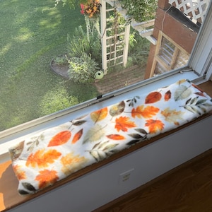 May include: A rectangular, plush fabric mat featuring a repeating pattern of autumn leaves in shades of orange, yellow, brown, and muted green against a white background, positioned horizontally along a wooden window sill.