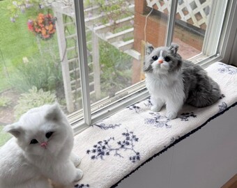 Puede incluir: Dos juguetes de peluche con forma de gato están en un alféizar. Uno es blanco, el otro gris y blanco. Están colocados sobre un cojín blanco con un estampado floral azul oscuro. La ventana da a un jardín.