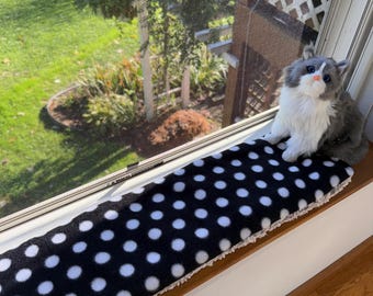 Puede incluir: Un juguete de peluche con forma de gato de pelaje gris y blanco, ojos azules y nariz rosa, sentado sobre un cojín rectangular largo para alféizar. El cojín presenta una parte superior de tela negra con lunares blancos y una base texturizada color crema.