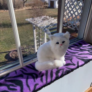 May include: A fluffy white cat figurine with pink nose and blue eyes sits on a purple and black zebra-striped window seat cushion. The cat is positioned in front of a window with a view of a sunny outdoor scene.