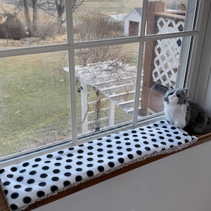 May include: A rectangular window perch mat featuring a white fabric base with a repeating pattern of black polka dots. The mat is positioned on a wooden windowsill next to a small, plush, grey and white cat figurine looking out the window.