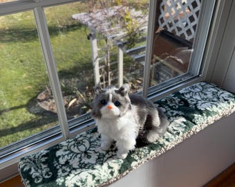 Puede incluir: Un juguete de peluche de gato gris y blanco con una nariz rosa, sentado en un cojín con estampado verde y blanco en un alféizar. La ventana da a un jardín soleado.