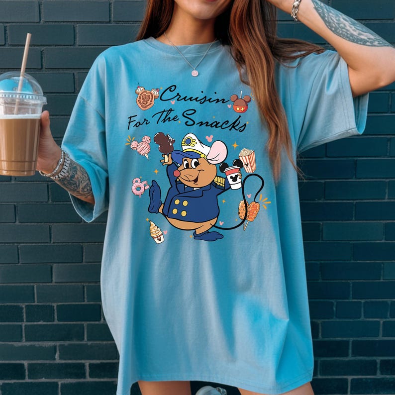May include: Light blue t-shirt with the text "Cruisin' For The Snacks" and a cartoon mouse character wearing a navy blue uniform. The mouse is holding ice cream, popcorn, and a coffee cup. The shirt has a relaxed fit.
