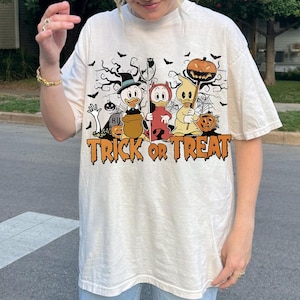 May include: Cream-colored t-shirt featuring a Halloween-themed graphic. The design includes three cartoon ducks dressed in costumes, a pumpkin, bats, and the words "TRICK OR TREAT" in orange. The shirt has short sleeves and a relaxed fit.