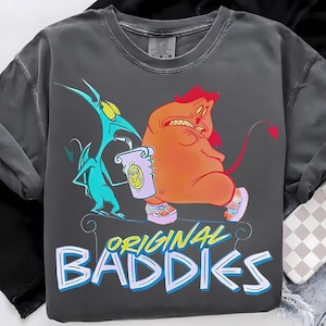 Peut inclure: T-shirt gris foncé avec une illustration de dessin animé de deux personnages. L'un est turquoise, tenant un parchemin, l'autre est orange avec des cheveux rouges. Le texte "ORIGINAL BADDIES" est en bleu et jaune.