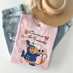 May include: A light pink t-shirt with the text "Cruisin' for the Snacks" and a cartoon mouse character holding a coffee cup and snacks. The shirt is folded on top of blue jeans and a tan hat.