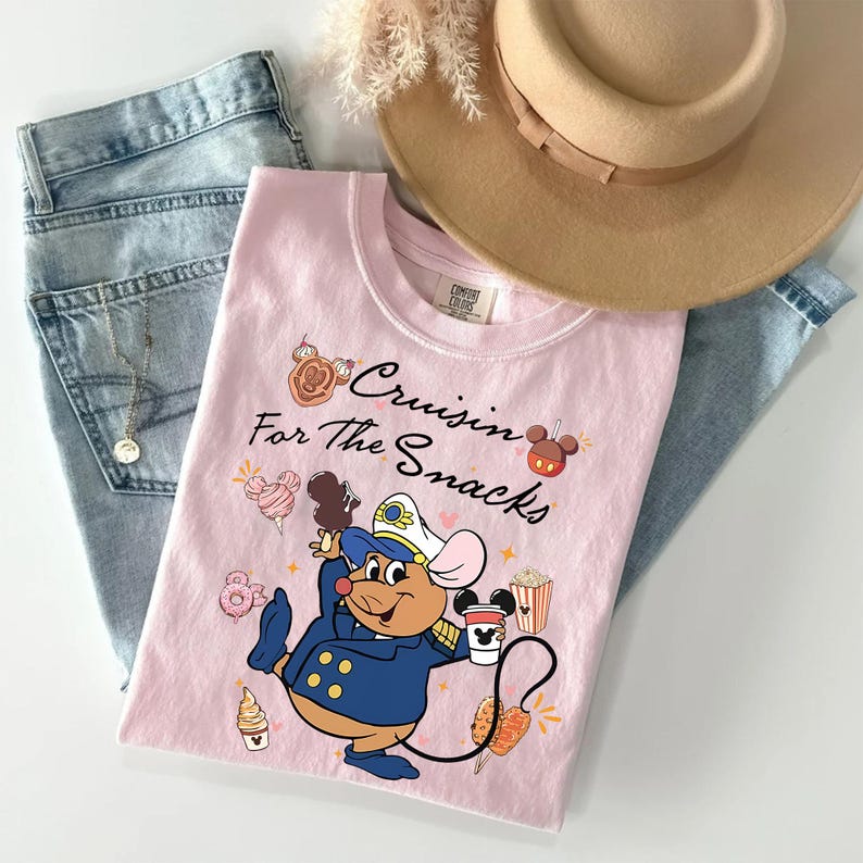May include: A light pink t-shirt with the text "Cruisin' for the Snacks" and a cartoon mouse character holding a coffee cup and snacks. The shirt is folded on top of blue jeans and a tan hat.