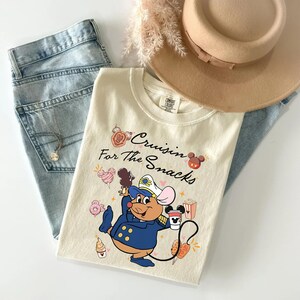 May include: Cream-colored t-shirt with the phrase "Cruisin' For The Snacks" and a cartoon mouse in a navy blue captain's uniform. The shirt is paired with light-wash jeans, a tan hat, and a necklace.