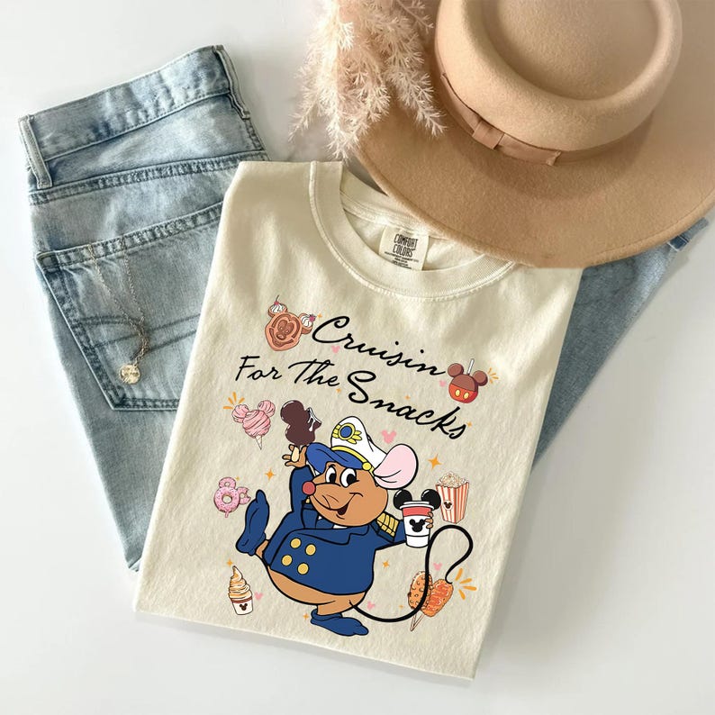 May include: Cream-colored t-shirt with the phrase "Cruisin' For The Snacks" and a cartoon mouse in a navy blue captain's uniform. The shirt is paired with light-wash jeans, a tan hat, and a necklace.