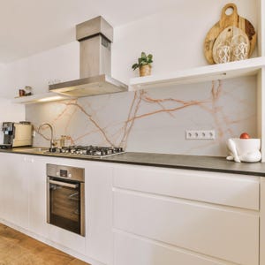 Op de afbeelding: Een moderne keuken met witte kasten, een roestvrijstalen afzuigkap en een ingebouwde oven. Het aanrechtblad is donkergrijs en de achterwand heeft een marmeren ontwerp met rode aderen. Een kleine plant staat op een plank.