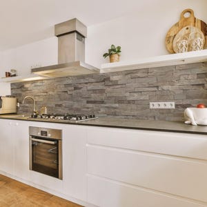 Può includere: Cucina moderna con mobili bianchi, una cappa in acciaio inox e un paraschizzi in pietra grigia. Sono visibili un forno a incasso, un piano cottura a gas e una macchina per il caffè. Taglieri in legno e una piccola pianta sono su una mensola.