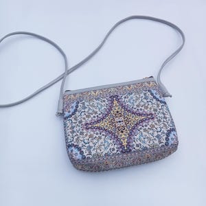 Op de afbeelding: Een rechthoekige crossbodytas met een grijze riem. De tas heeft een gedetailleerd, kleurrijk patroon dat doet denken aan een Perzisch tapijt, met een centraal medaillonontwerp in paars, goud en blauw. De tas heeft een ritssluiting.