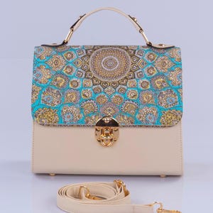 Op de afbeelding: Een crèmekleurige satchel-handtas met een gouden sluiting en een afneembare schouderband. De voorflap heeft een kleurrijk patroon met blauwe, gouden en witte accenten.