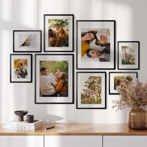 Könnte beinhalten: Eine Anordnung von gerahmten Familienfotos in verschiedenen Größen an einer weißen Wand. Die schwarzen Rahmen enthalten Farbfotos von Menschen. Auf einem Holztisch stehen dekorative Gegenstände.