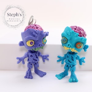May include: Two zombie figurines, one purple and one teal, with exposed brains and open mouths. The zombies have yellow eyes and green teeth. The figurines are standing on a white surface. The text "Steph's Magical Menagerie" is in a circle in the upper left corner.