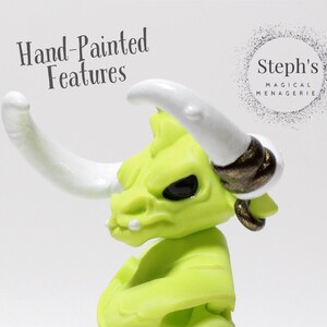 May include: A hand-painted, lime green figurine of a horned creature with white horns and black eyes. The figurine has a gold earring and a small white tooth. The text "Hand-Painted Features" is visible. The Steph's Magical Menagerie logo is also visible.