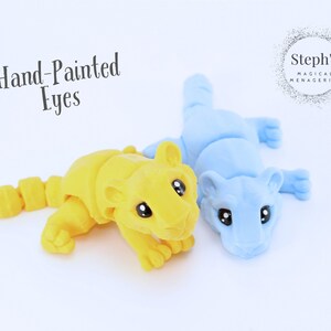 May include: Two hand-painted animal figurines, one yellow and one blue, with large eyes. The figurines are posed in a playful, lying down position. The text "Hand-Painted Eyes" is visible above the figurines. The text "Steph's Magical Menagerie" is visible in the upper right corner of the image.