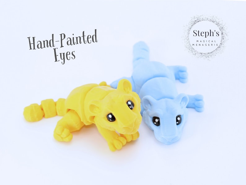 May include: Two hand-painted animal figurines, one yellow and one blue, with large eyes. The figurines are posed in a playful, lying down position. The text "Hand-Painted Eyes" is visible above the figurines. The text "Steph's Magical Menagerie" is visible in the upper right corner of the image.