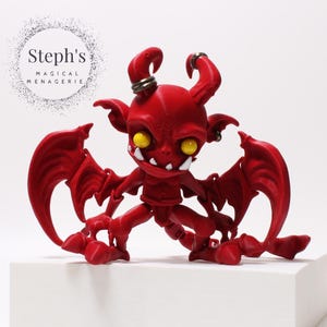 Op de afbeelding: Een rode figuur van een duivelachtig wezen met gele ogen, kleine hoorns en grote vleugels. De figuur heeft een dreigende uitdrukking en staat op een wit blok. De tekst "Steph's MAGICAL MENAGERIE" staat in een cirkel op de achtergrond.