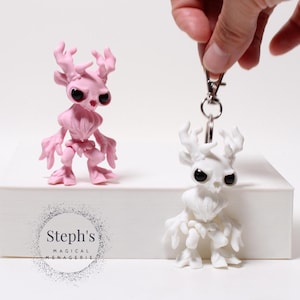 Puede incluir: Dos figuras caprichosas, una rosa y otra blanca, con rasgos de cuernos y grandes ojos negros. La figura blanca está unida a un llavero. La figura rosa está de pie sobre una caja blanca. El texto "Steph's Magical Menagerie" es visible.