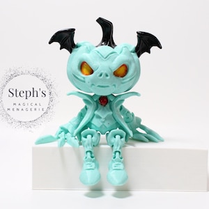 Könnte beinhalten: Hellblaue Figur einer Kreatur mit Fledermausflügeln, Hörnern und leuchtend orangefarbenen Augen. Die Figur hat einen roten Edelstein auf der Brust und sitzt auf einem weißen Block. Der Text "Steph's Magical Menagerie" ist im Hintergrund.