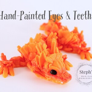 Pu&ograve; includere: Due figurine di drago arancioni e gialle dipinte a mano con squame dettagliate e occhi e denti dipinti a mano. Le figurine sono su uno sfondo bianco. Steph's Magical Menagerie.