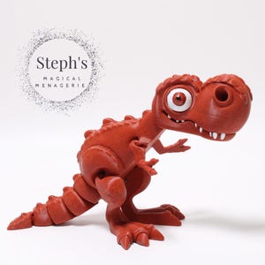 May include: A red, cartoon-style dinosaur toy with a large, white eye. The dinosaur is standing on a white surface. Steph's Magical Menagerie is written in a circle above the dinosaur.