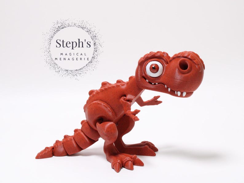 May include: A red, cartoon-style dinosaur toy with a large, white eye. The dinosaur is standing on a white surface. Steph's Magical Menagerie is written in a circle above the dinosaur.