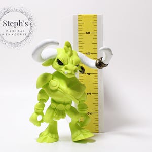 May include: A lime green figurine of a bull-like creature with white horns and a gold ring on one horn. The figurine is next to a yellow ruler. The text "Steph's Magical Menagerie" is in a circle in the background.
