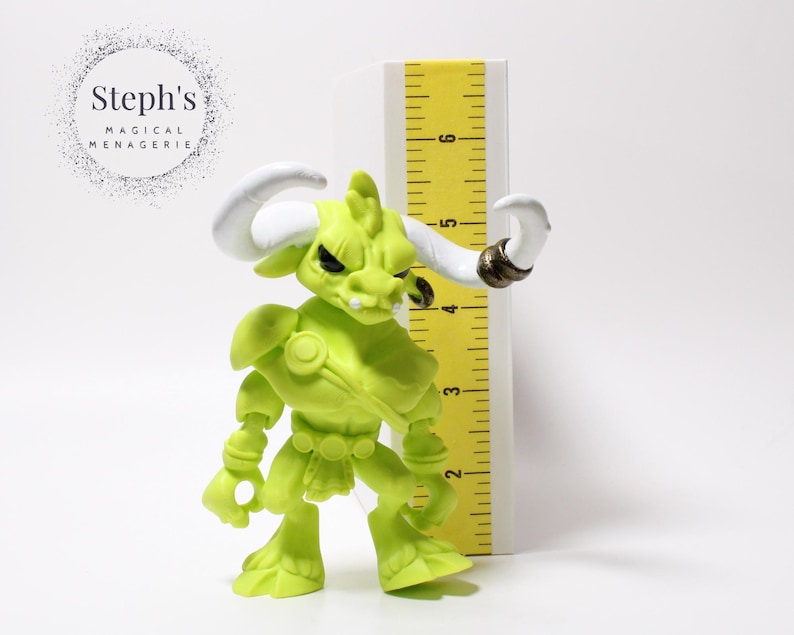 May include: A lime green figurine of a bull-like creature with white horns and a gold ring on one horn. The figurine is next to a yellow ruler. The text "Steph's Magical Menagerie" is in a circle in the background.