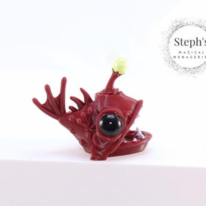 May include: A red anglerfish figurine with a bioluminescent lure on its head. The figurine has a large, black eye and a wide, open mouth with sharp teeth.