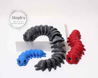 Puede incluir: Tres juguetes de oruga articulados en azul, negro y rojo. La oruga negra está curvada, mientras que las azules y rojas son rectas. Cada una tiene un cuerpo segmentado y ojos grandes y redondos. El logo de Steph's Magical Menagerie está arriba a la izquierda.