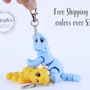 May include: Two small, articulated, 3D printed animal figures. The blue figure is on top of the yellow figure. Both figures are attached to a silver key chain.