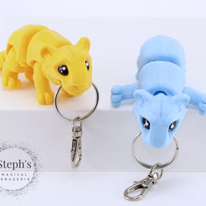 May include: Two small, articulated animal keychains. One is yellow and the other is light blue. Both have black eyes and are posed in a playful manner. The keychains are attached to silver key rings with a silver clasp.