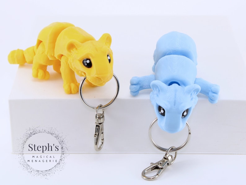 May include: Two small, articulated animal keychains. One is yellow and the other is light blue. Both have black eyes and are posed in a playful manner. The keychains are attached to silver key rings with a silver clasp.
