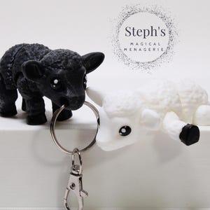 May include: Black and white sheep keychain charms attached to a silver key ring. The black sheep is on the left and the white sheep is on the right. The keychain is on a white surface. Steph's Magical Menagerie is written in a circle above the sheep.
