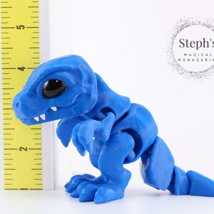 May include: A blue articulated dinosaur figurine with large black eyes and white teeth. The figurine is approximately 4 inches tall and is positioned next to a yellow ruler. The figurine is made of a smooth material and has a segmented body.