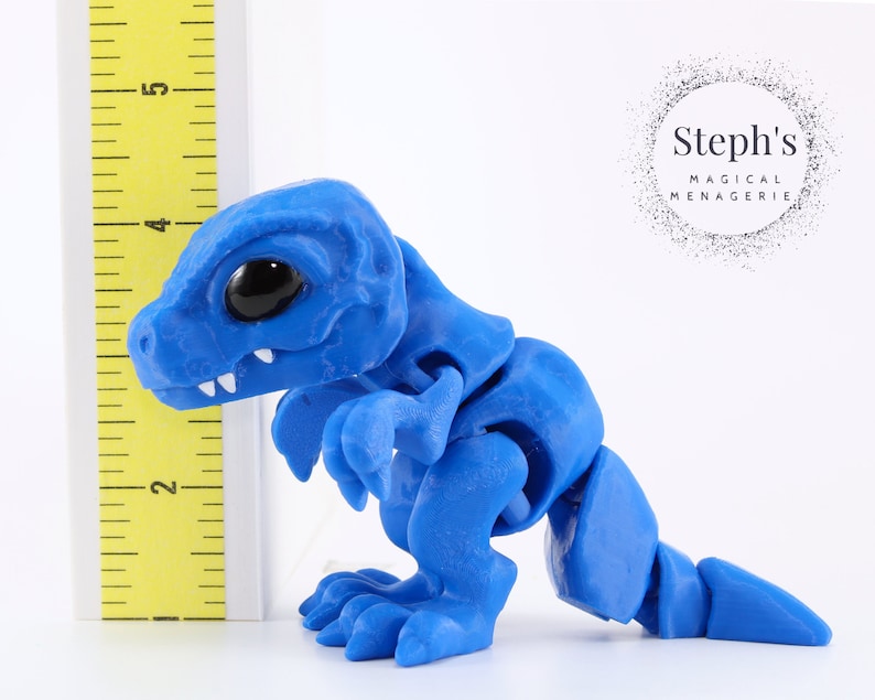 May include: A blue articulated dinosaur figurine with large black eyes and white teeth. The figurine is approximately 4 inches tall and is positioned next to a yellow ruler. The figurine is made of a smooth material and has a segmented body.