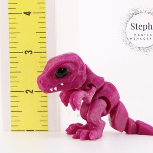 May include: A magenta articulated dinosaur figurine with black eyes and white teeth. The figurine is next to a yellow ruler, approximately 3 inches tall. The product is from Steph's Magical Menagerie.