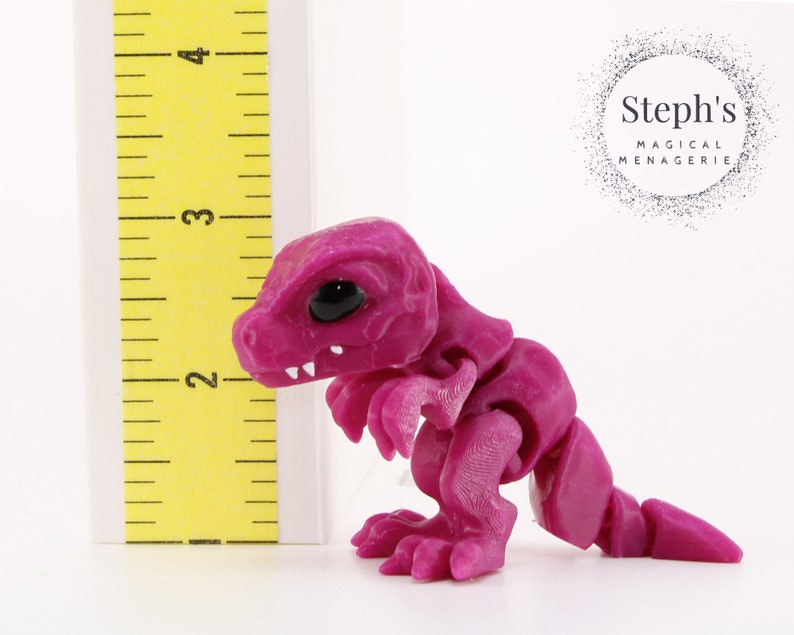 May include: A magenta articulated dinosaur figurine with black eyes and white teeth. The figurine is next to a yellow ruler, approximately 3 inches tall. The product is from Steph's Magical Menagerie.