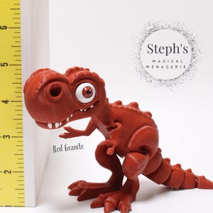 May include: Red granite dinosaur figurine with large eyes and a toothy grin. The figurine is standing on a white surface with a yellow ruler behind it. The text "Steph's Magical Menagerie" is visible in the background.