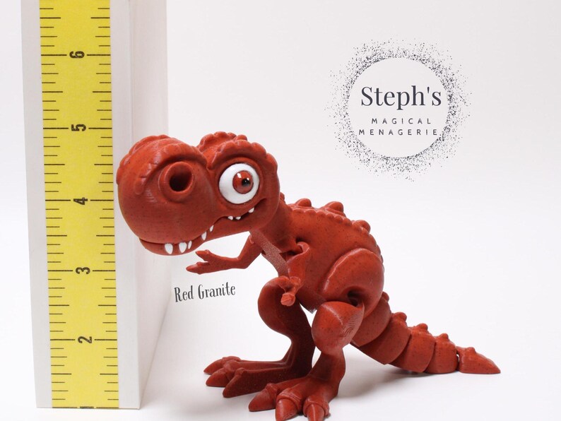 May include: Red granite dinosaur figurine with large eyes and a toothy grin. The figurine is standing on a white surface with a yellow ruler behind it. The text "Steph's Magical Menagerie" is visible in the background.