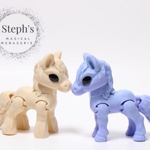 Puede incluir: Dos caballos de juguete articulados, uno crema y otro azul lavanda, se enfrentan. El caballo crema tiene crin y cola claras, mientras que el caballo azul lavanda tiene crin y cola más oscuras. El texto "Steph's MAGICAL MENAGERIE" está encima.