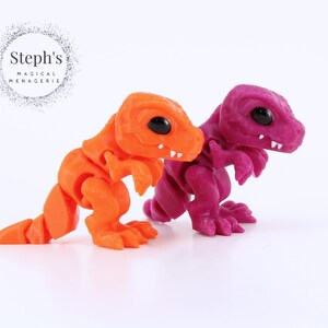 May include: Two articulated dinosaur figurines. One is bright orange, the other is a deep magenta. Both have black eyes and white teeth. The figurines are on a white background. The text "Steph's Magical Menagerie" is in the upper left corner.