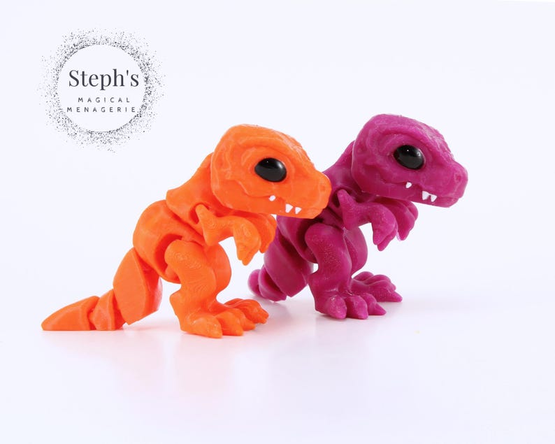 May include: Two articulated dinosaur figurines. One is bright orange, the other is a deep magenta. Both have black eyes and white teeth. The figurines are on a white background. The text "Steph's Magical Menagerie" is in the upper left corner.