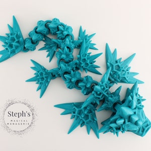 May include: A turquoise blue, 3D printed, articulated dragon sculpture with a spiky, crystalline design. The dragon is posed in a crawling position with its head raised.