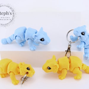 May include: Four articulated animal keychains, two are yellow and two are light blue. The keychains are posed in a playful manner. Steph's Magical Menagerie is printed on the background.